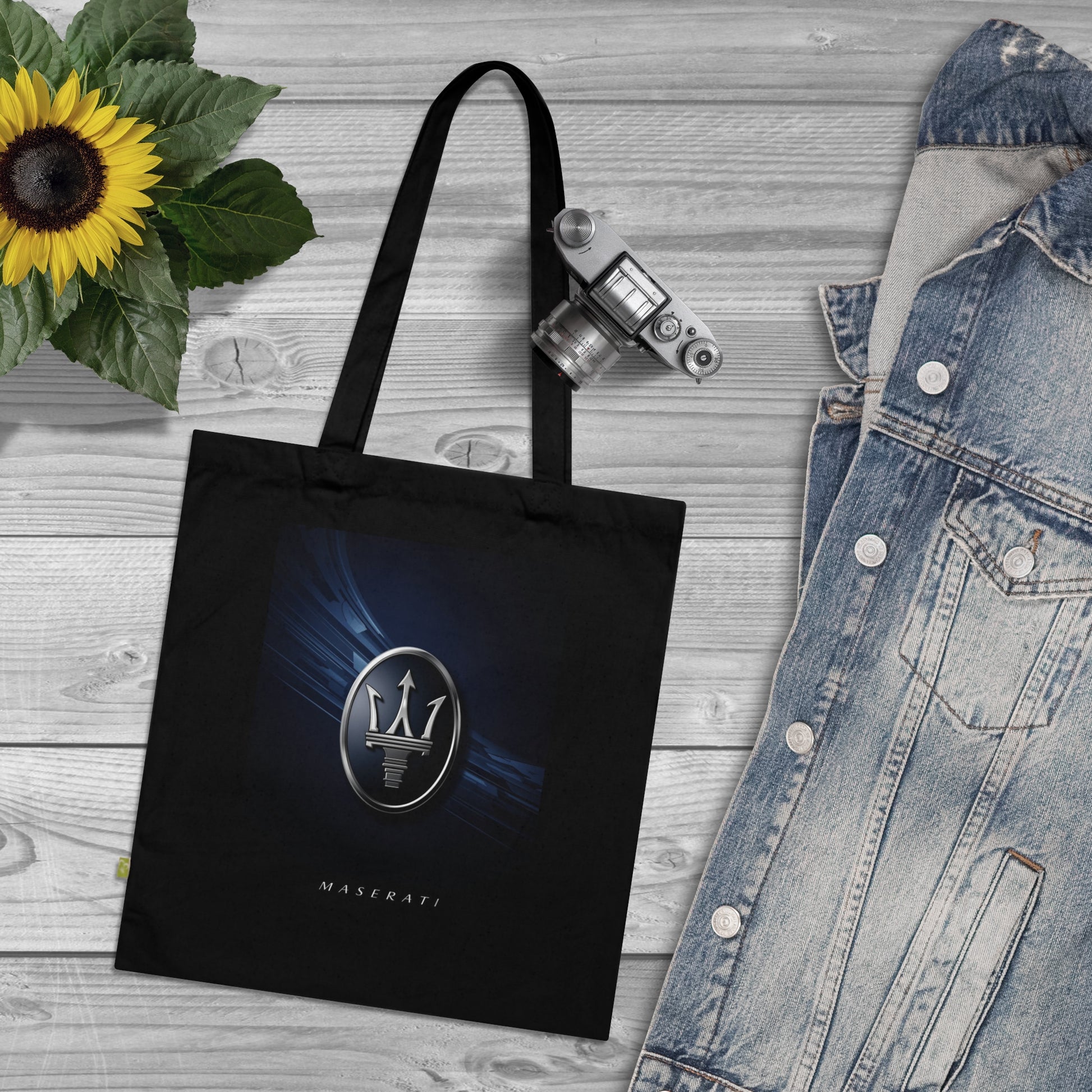 Black tote bag with Maserati logo on a wooden surface with denim jacket and sunflower.