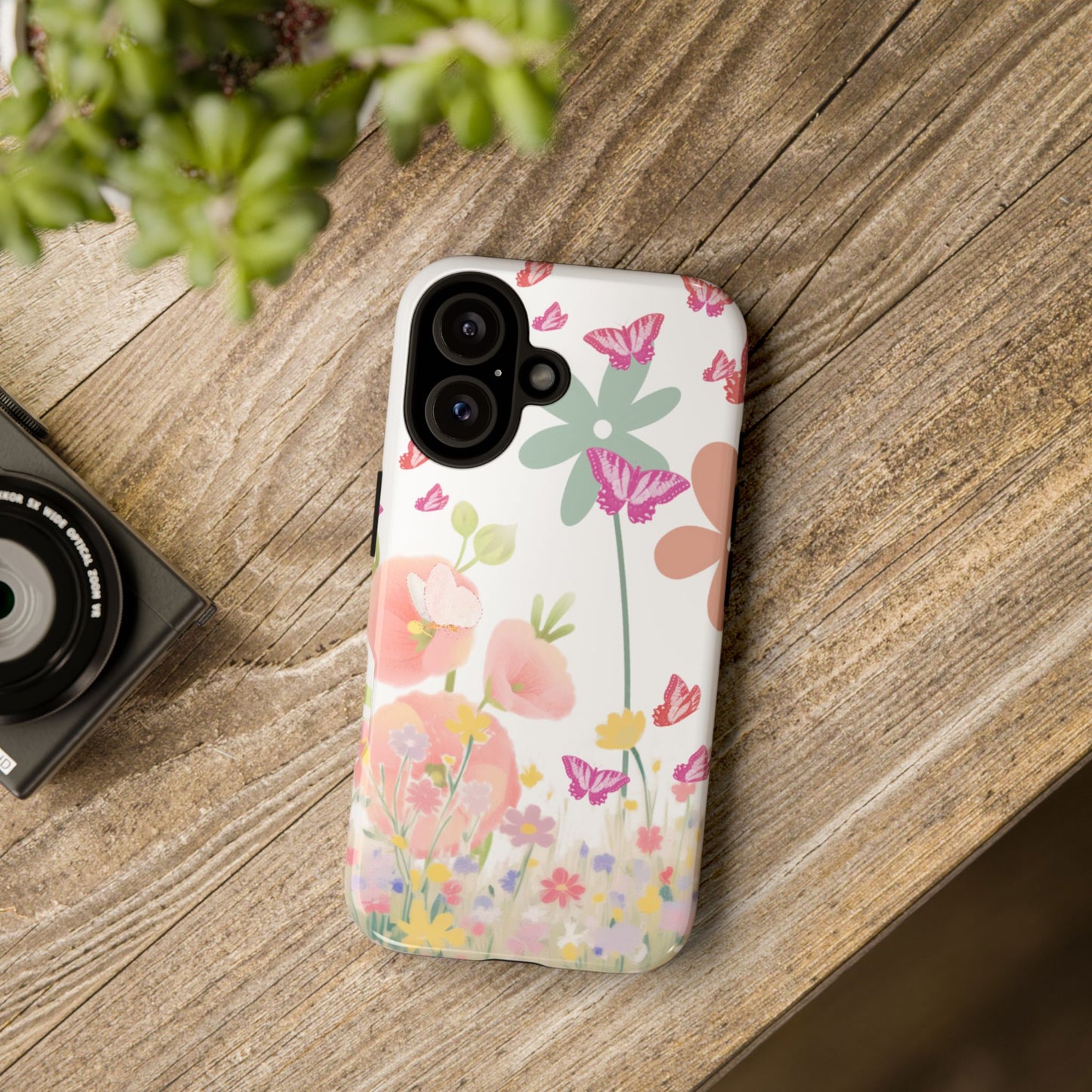 Phone case with floral design on a wooden surface next to a camera and plant