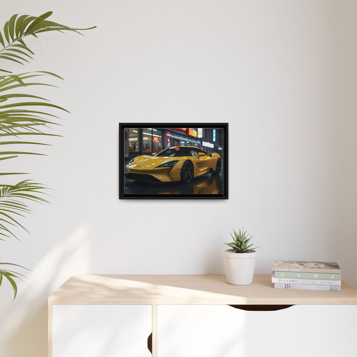 Framed picture of a yellow sports car on a wall above a desk with a plant and books.