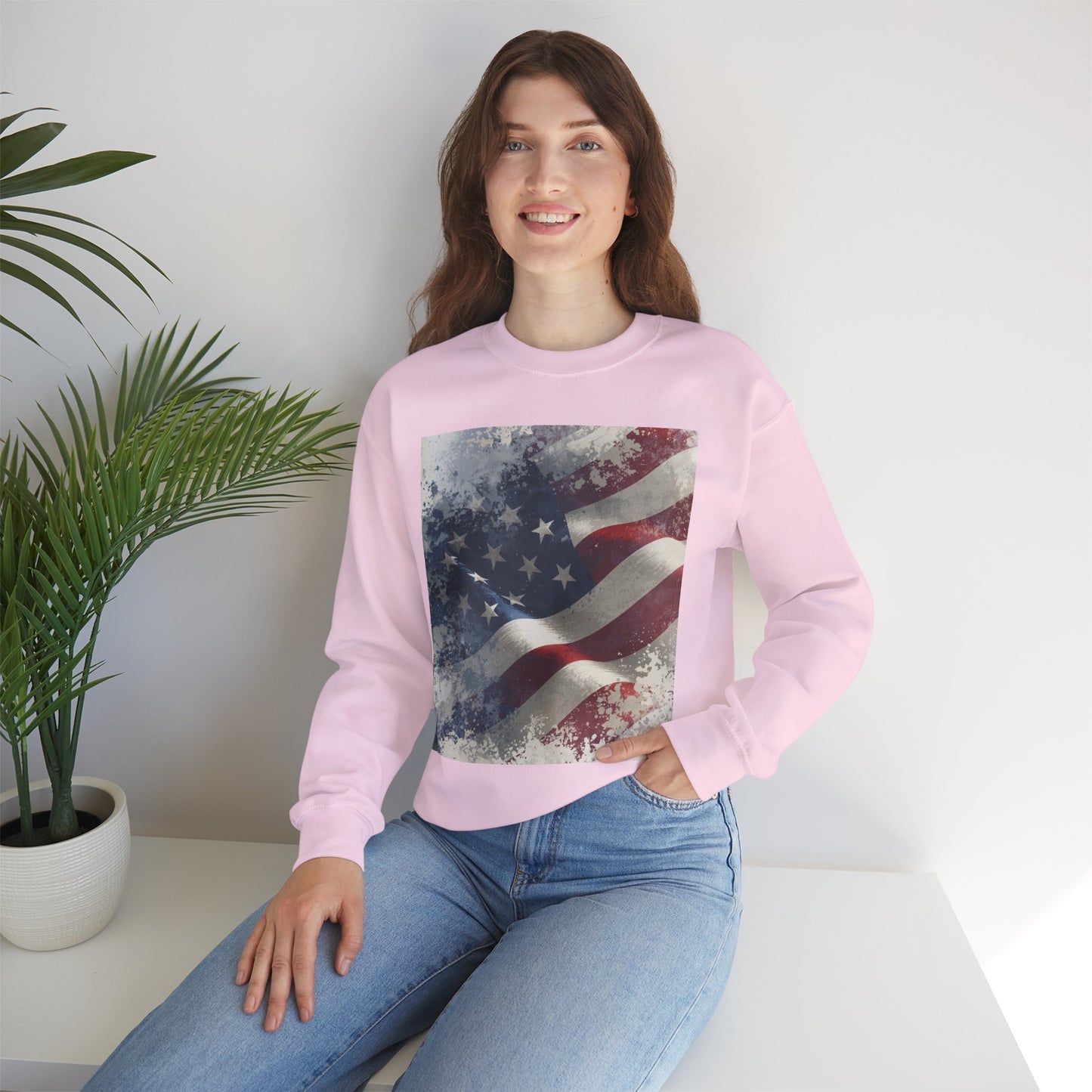 Woman wearing a pink sweatshirt with a vintage American flag design, sitting on a white surface with a plant in the background.