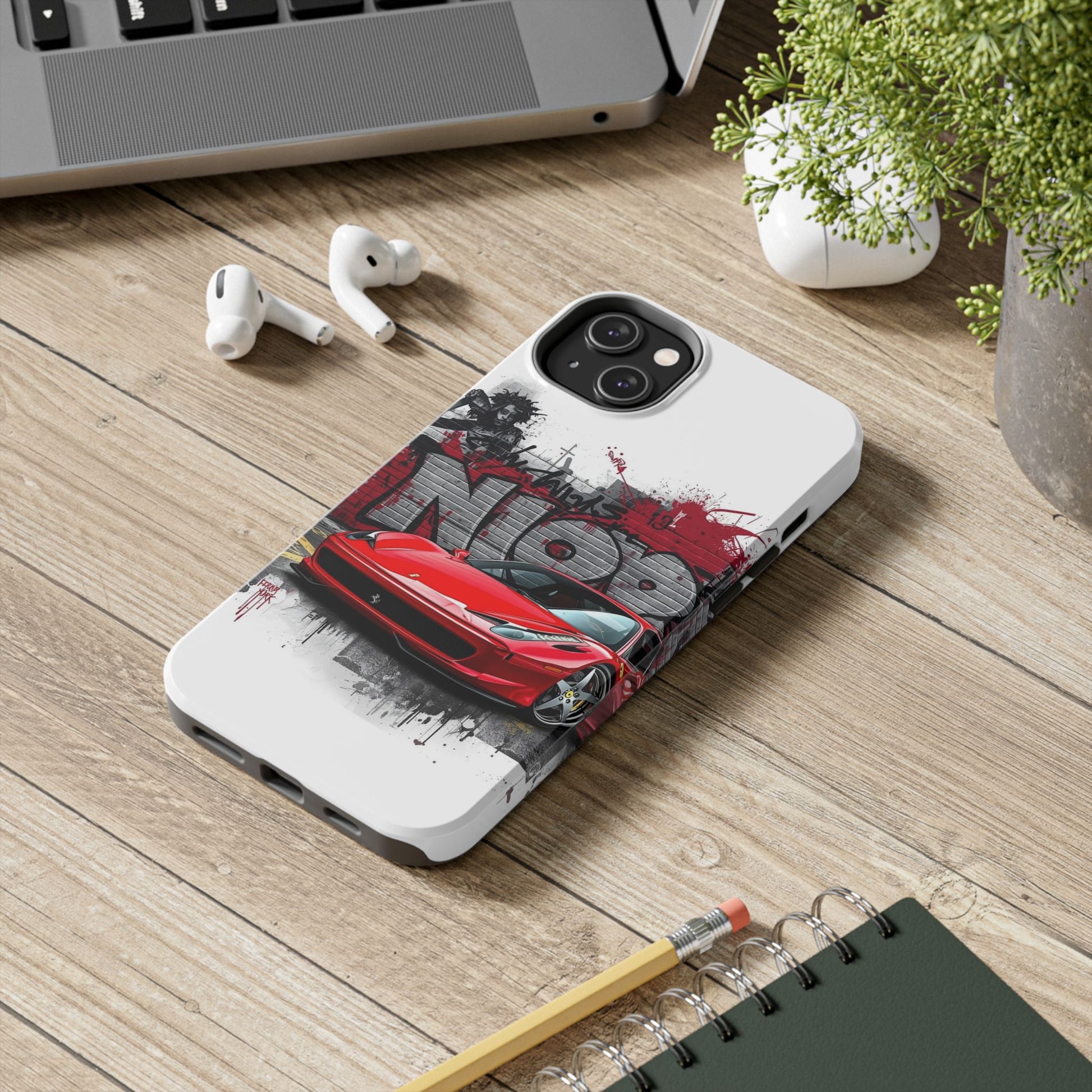 Phone case with red car design on a wooden desk with laptop, earbuds, and notebook.