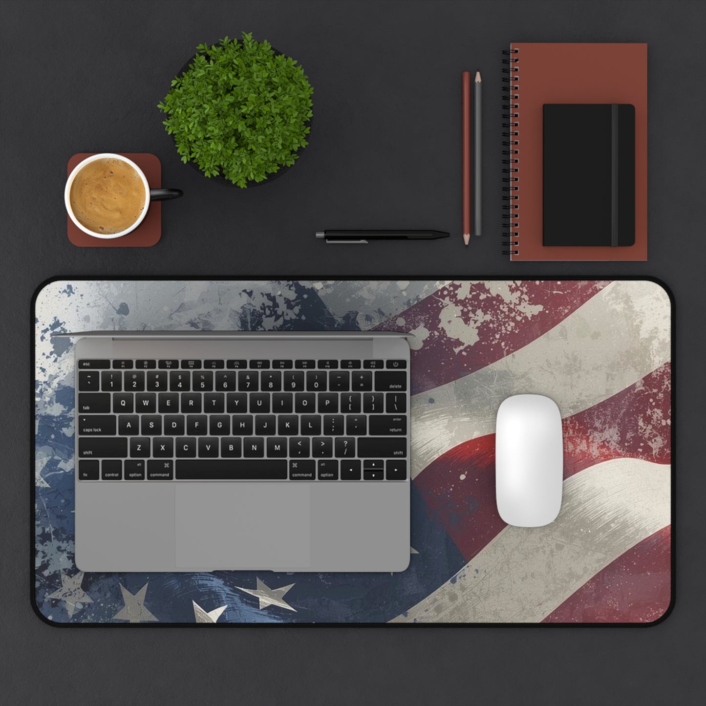 Laptop on a desk with an American flag design mouse pad desk mat, cup, plant, and office supplies.