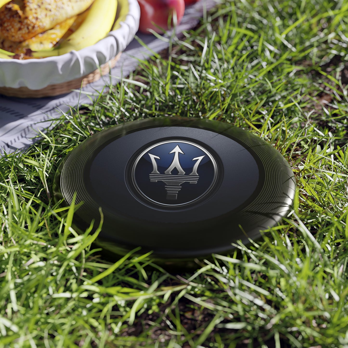Black Maserati logo on a grassy surface with a picnic setup in the background.