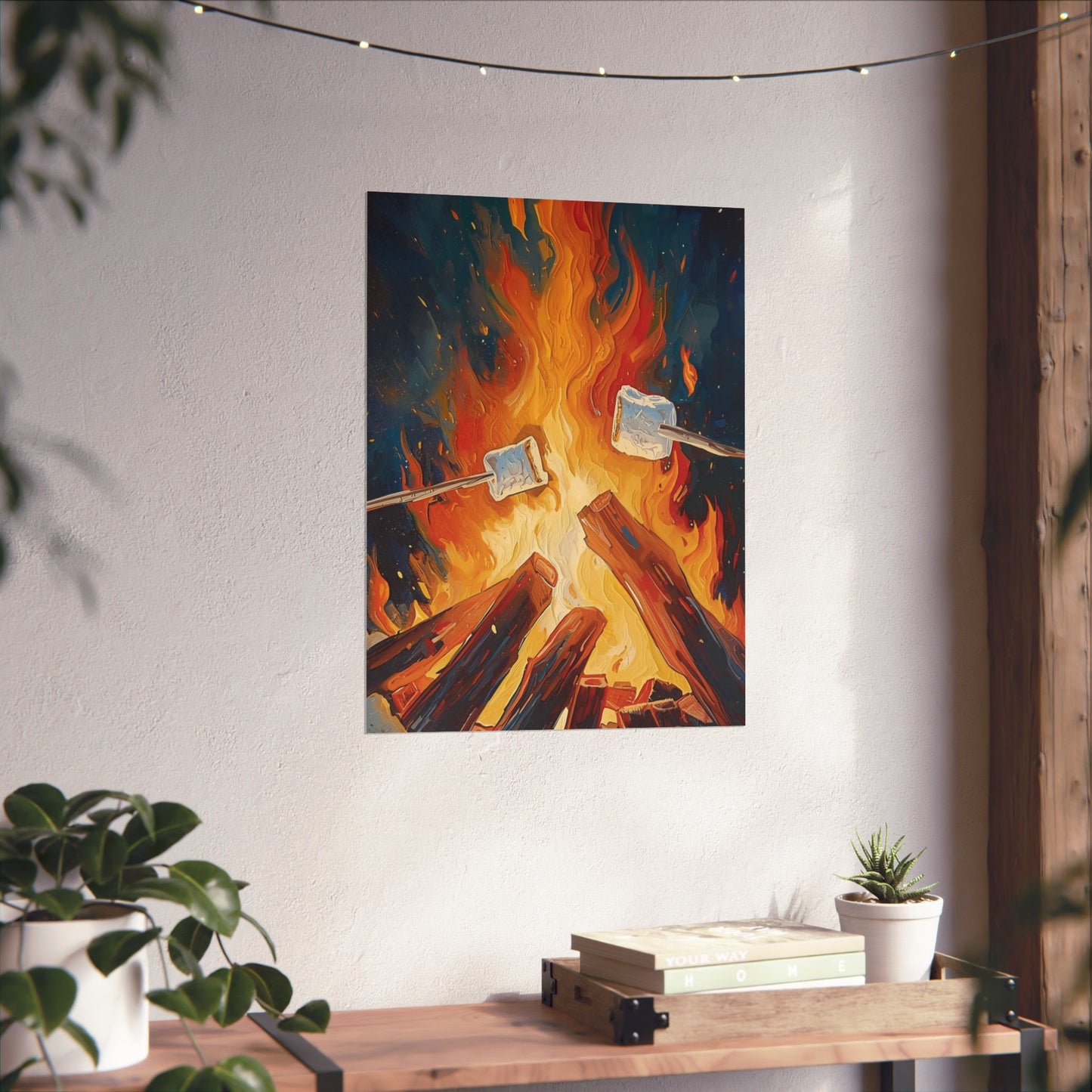 Art print of marshmallows roasting over a campfire on a wall above a wooden shelf with plants and books.