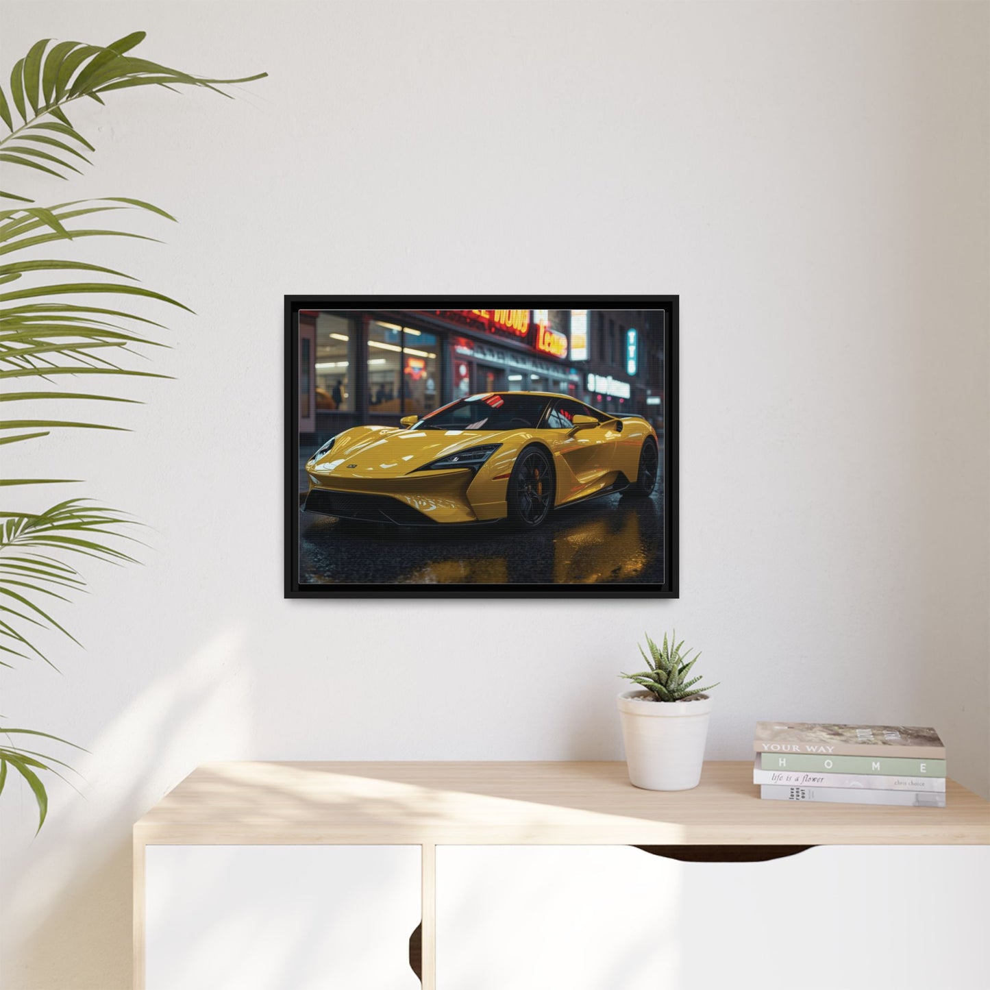 Framed artwork of a yellow sports car on a wall above a desk with a plant and books.